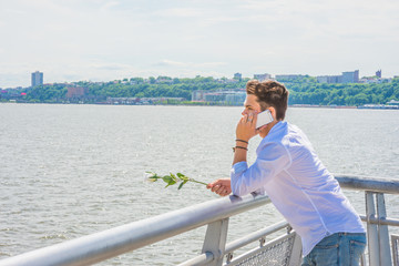 Wearing white shirt, jeans, holding white rose, a young guy standing by Hudson River in New York, opposite New Jersey, listening, talking on mobile phone. Concept of looking for love, friendship..