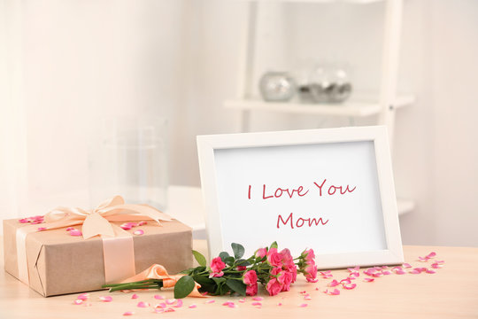 Frame with text I LOVE YOU MOM, gift box and flowers on table