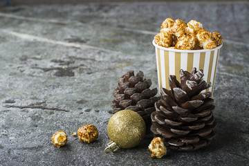 Appetizing golden caramel popcorn in paper striped cups in the New Year's interior with fir cones, New Year's golden balls on a gray stone modern background. Selective focus.