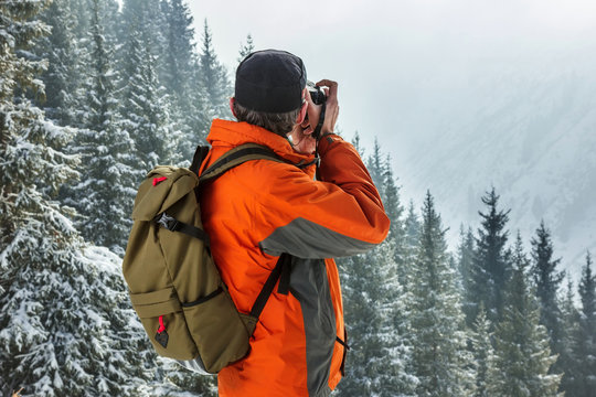A man photographes a winter landscape. Against the background of mountains and pines. Pictured from the back