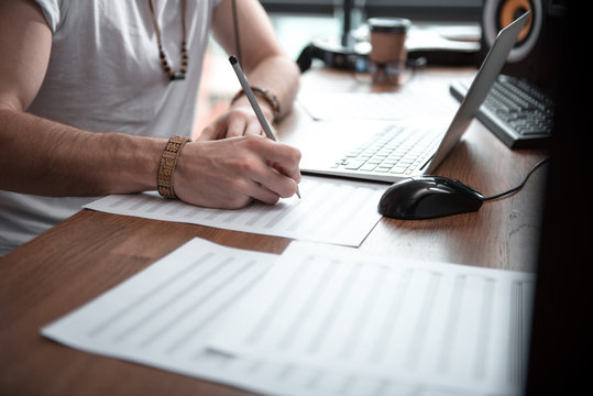 Close up of male hands drawing musical notes on paper. Man is sitting at desk near laptop
