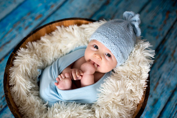 Little baby boy with knitted hat in a basket, happily smiling - obrazy, fototapety, plakaty