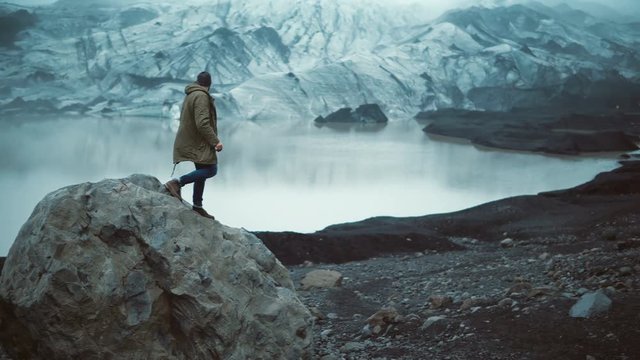 Young handsome man sitting on the rock in ice lagoon and looking on glaciers in Iceland. Tourist stand up and going away