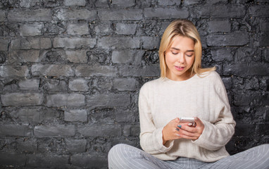 Beautiful hipster girl chatting in social network on mobile phone, sitting against brick wall with copy space for advertising text. Charming blonde woman student typing text on portable cellphone