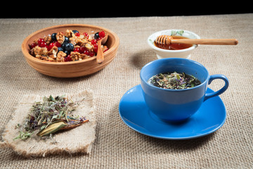  real herbal tea in a blue Cup filled on a napkin, and honey in a bowl of honeycomb in a wooden bowl with red currants and blueberries or blueberry on a background of a linen tablecloth