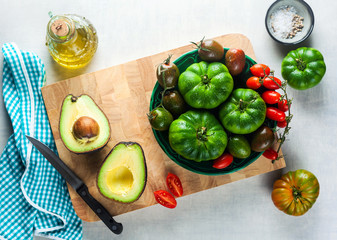 ingredients for salad from different types of tomato, avocado and arugula on a white stone table. cutting board, olive oil and condiments. healthy food, summer or spring menu