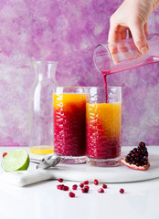 pouring grenadine into two glasses of alcoholic cocktail with orange liqueur, freshly squeezed pineapple and pomegranate juice and lime and ice on the table.
