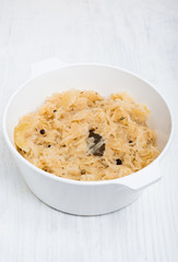 Traditional German Sauerkraut cooked with white wine, apples, onions, bay leaves, peppercorns, juniper berries, caraway and allspice in white pot on white wooden background