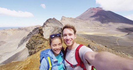 Young couple hiking taking selfie video at hike Tongariro Alpine Crossing. Smiling healthy backpackers photographing themselves in mountains. Beautiful landscape in Tongariro National Park New Zealand - Powered by Adobe