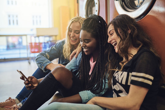 Diverse Young Girlfriends Using A Cellphone Together At The Laundromat