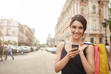 Portrait of cheerful attractive young caucasian woman with dark hair in black dress smiling in camera with teeth, holding shopping bags and smartphone in hands, catting with friend. Soft focus