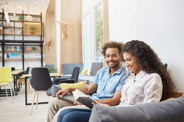 Close up of two young people in casual clothes sitting on sofa in modern library. Girl reading to book, getting ready for universit lecture. Guy looking in camera with happy face expression, holding