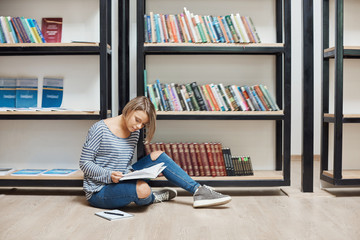 Portrait of young good-looking student girl with short blonde hair in casual stylish clothes sitting on foor in modern library near shelfs, reading favourite books, spending weekend in cosy atmosphere