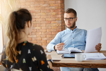 Business concept. Mature bearded serious male company director sitting in office in front of dark-haired girl who came on job interview. Looking through papers, talking about work experience.