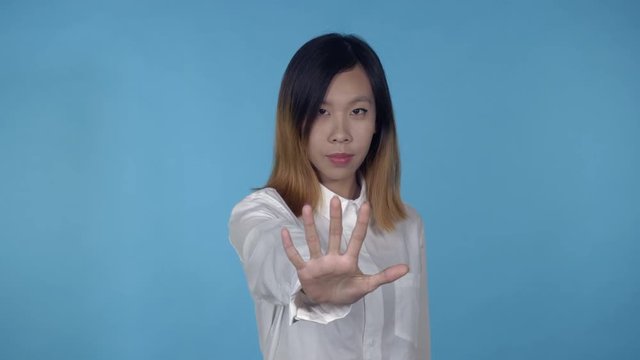 young asian woman posing showing hand gesture stopping on blue background in studio. attractive millennial girl wearing white casual shirt looking at the camera smiling