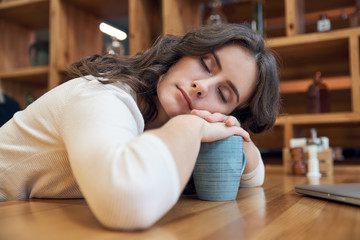 Obraz na płótnie Canvas Attractive long-haired girl tiredly falls asleep at a table in a cafe. The concept of morning awakening, tiredness, insomnia, long wait, not the vigor