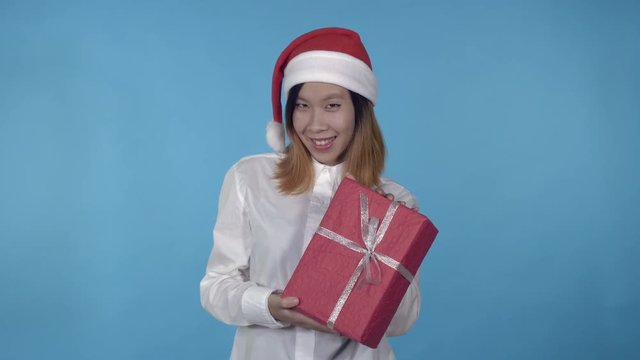young asian woman posing wearing santa claus hat showing red box with present on blue background in studio. attractive millennial girl wearing white casual shirt looking at the camera laughing