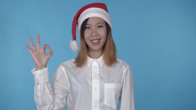 young asian woman posing wearing santa claus hat showing symbol hand gesture ok on blue background in studio. attractive millennial girl wearing white casual shirt looking at the camera laughing