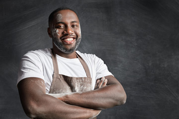 Portrait of cheerful dark skinned black male chef has pleasant smile, keeps hands crossed, being glad to be praised by clients and recieve culinary reward, wears dirty apron, isolated over black wall