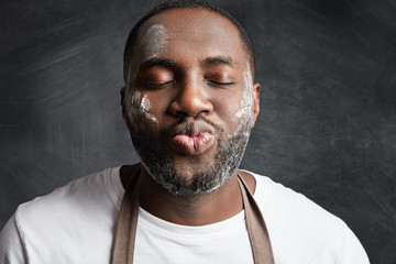 Close up portrait of dark skinned male being satisfied after baking something delicious, roundes lips and closes eyes in enjoyment, has dirty face with flour poses against black chalk background.