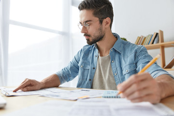 Serious concentrated male worker checks and analyzes information from business report, writes necessary figures in notebook, compares prices from different departments, engaged in working process