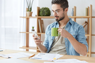 Indoor shot of handsome bearded man dressed casually, holds smart phone and types messages to...