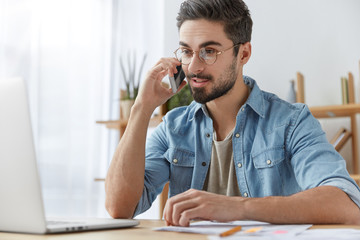 Fototapeta premium Portrait of confident bearded male freelancer manages all problems as uses modern gadgets, speaks over smart phone with colleague and reads information on laptop computer, browses in internet