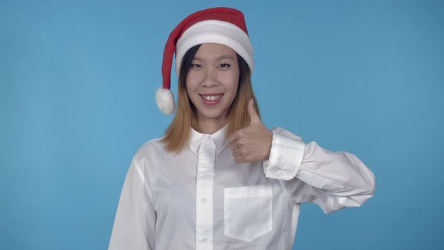 young asian woman posing wearing santa claus hat showing symbol hand gesture like on blue background in studio. attractive millennial girl wearing white casual shirt looking at the camera laughing
