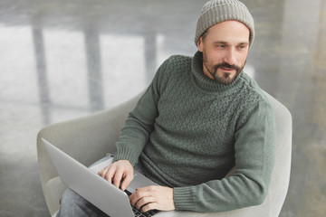 Thoughtful middle aged man with thick beard and mustache, wears warm knitted clothes, reads news in network via modern laptop computer, searches information in internet, looks thoughtfully aside