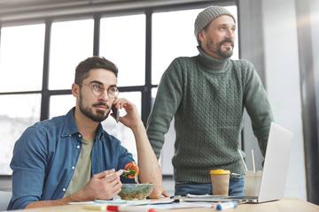 Attractive bearded man wears stylish clothes, holds smart phone as has phone conversation, eats fresh vegetable salad, has break after working with documents and his smiling partner stands near