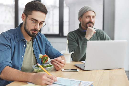 Two coworkers works together at common project, dressed casually. Bearded young male works on documents, has fast snack, eats healthy salad and his partner in background uses laptop computer