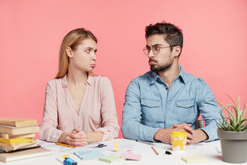 Discontent upset male and female student realize that tomorrow is exam, looks at each other with upset expressions, curve lips, sit at workplace, surrounded with books and papers, prepare actively