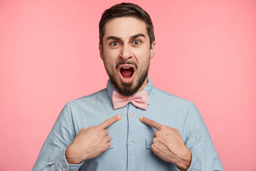 Displeased indignant male indicates at his new shirt, shows spot, going on meeting, being puzzled as it is dirty. Annoyed bearded man points at himself, discontent to be chosen leader of team