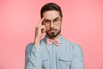 Waist up portrait of thoughtful serious stylish man keeps finger on temple, tries to concentrate and make important decision, poses agianst pink studio background. Focused male enterpreneur.