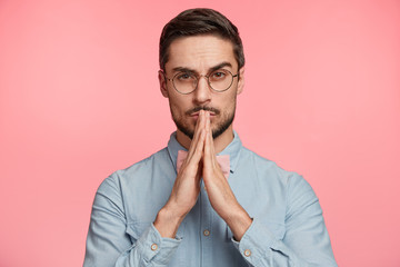 Hopeful serious faithful young male with trendy hairdo, beard and mustache, keeps hands in praying gesture, worries before important event, isolated over pink background. People and wish concept