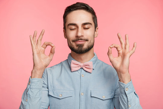 Concentrated bearded man wears formal shirt and tie bow, makes ok sign, closes eyes, shows approval or tries to relax, poses against pink background, meditates indoor. People, body language concept