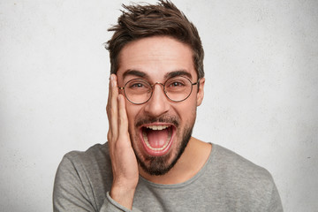 Positive excited fashionable male has trendy hairdo, wears round spectacles, keeps hand on cheek, laughs as hears funny joke or story, expresses positiveness, isolated over white background.