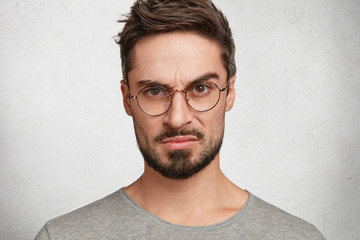 Obraz premium Close up portrait of bearded serious man with strict expression, has hazel eyes, looks directly into camera with confident look, raises eyebrow. Male business owner listens to investment plan