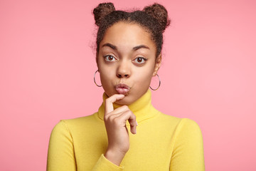 Beautiful young female model has two hair buns, wears casual clothes, rounds lips, being surprised or looks at something with enjoyment, isolated over pink background. People, body language concept