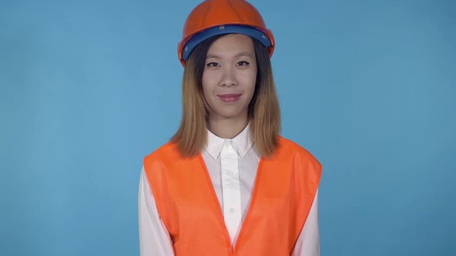young asian woman posing on blue background in studio. attractive millennial girl wearing white casual shirt and orange hard hat and vest looking at the camera smiling