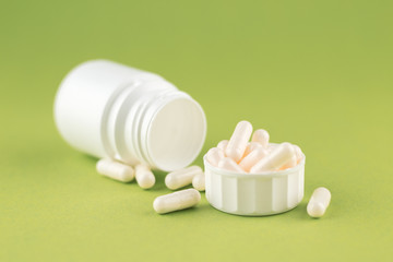 Close up white pill bottle with spilled out pills and capsules in cap on olive yellow background with copy space. Focus on foreground, soft bokeh. Pharmacy drugstore concept