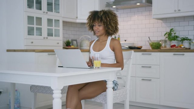 Pretty ethnic woman in home clothing sitting at table in kitchen having glass of fresh juice and watching laptop chatting online.