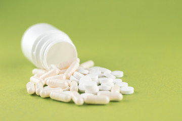 Close up white pill bottle with spilled out pills and capsules on olive yellow background with copy space. Focus on foreground, soft bokeh. Pharmacy drugstore concept