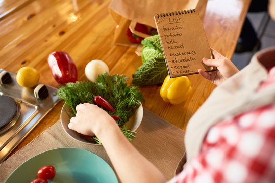 Close-up shot of unrecognizable woman wearing checked shirt studying list of ingredients necessary for healthy dish while standing at kitchen table
