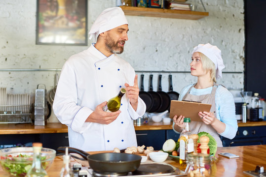 Interactive cooking workshop at full speed: handsome middle-aged chef giving instructions to pretty young woman while she taking necessary notes, interior of modern kitchen on background