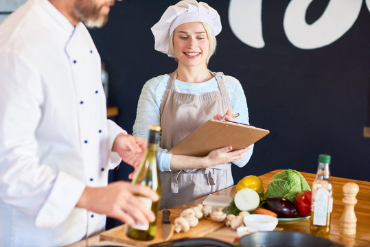 Portrait of smiling young assistant wearing apron taking necessary notes while bearded chef showing her how to prepare dish correctly, interior of modern restaurant kitchen on background