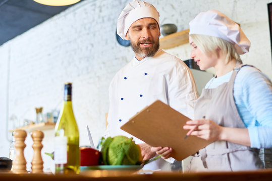 Handsome bearded chef wearing uniform sharing culinary secrets with pretty young assistant while preparing dish together, interior of modern restaurant kitchen on background