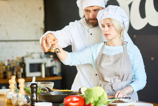 Concentrated middle-aged chef and pretty young woman wearing apron pouring olive oil into frying pen together while preparing dish at cooking workshop
