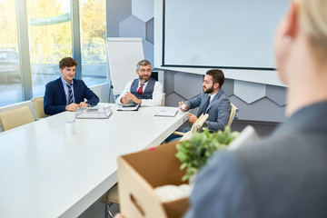 Group of hard-working white collar workers in classical suits sitting at boardroom table and analyzing results of accomplished work, unrecognizable female colleague holding cardboard box in hands