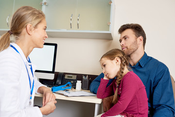 Fototapeta premium Portrait of smiling young pediatrician talking to man about health of his little girl, all sitting at desk in office